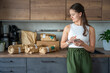 © dtatiana - Young woman checks grocery delivery using a mobile app while standing in the kitchen. Concept of app-based meal kit subscription and digital grocery planning for home cooking.