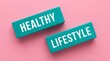 © Mary Patricia - Two teal colored wooden blocks with white text reading HEALTHY and LIFESTYLE are placed on a pink background wellbeing wellness motivation inspiration conceptual words messages quotes