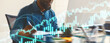 © Prostock-studio - A business professional is focused on creating graphs and analyzing data at a sleek desk in a contemporary office. The individual uses a notepad and pen while surrounded by digital graphics.