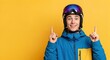 © MT - Happy young Caucasian man in blue and yellow ski jacket and helmet with goggles, pointing both index fingers upwards on a vibrant yellow studio background, ready for winter sports or advertising.