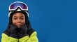 © MT - Smiling young African American boy in full ski gear, wearing a white helmet and purple goggles, posing cheerfully with hands under his chin against a vibrant blue studio background.
