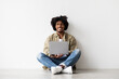 © Prostock-studio - A smiling African American man sits comfortably on the floor near a white wall, typing on his laptop. He enjoys his distance job, radiating positivity and relaxation in his workspace.