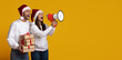 © Prostock-studio - A cheerful couple wearing Santa hats stands against a bright yellow background. One person holds wrapped gifts while the other joyfully uses a megaphone to spread holiday cheer.