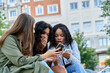 © Oscar - Three young women sharing exciting news on smartphone in urban setting