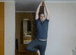 © Siniehina - Plus-size man practicing yoga at home on a mat, stretching to improve flexibility, joint mobility and posture. Concept of home fitness, rehabilitation, wellness and mindful movement.