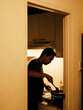 © Skyfe - Moody amber-lit doorway-framed portrait of man cooking on blue gas flame in small kitchen, silhouette, left copy space — home cooking editorial
