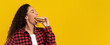 © Prostock-studio - A young woman with curly hair takes a big bite of a tasty hamburger while standing in front of a vibrant yellow backdrop. She is dressed casually in a plaid shirt, and it is likely midday.