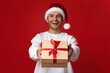 © Prostock-studio - A man wearing a Santa hat presents a festive gift box with a red ribbon. He is smiling widely, embodying holiday cheer in front of a bright red backdrop.