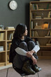 © Mediaphotos - Teenage Asian girl sitting on chair with knees drawn to chest, looking sideways during group therapy session with bookshelves, hands clasped around legs, thoughtful expression