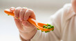 © Mirivox - Close-up of child's hand holding orange spoon with green peas, symbolizing infant feeding, nutrition, or early childhood food introduction