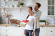 © BestForYou - Woman in a white long-sleeve shirt holding a red gift box with a white ribbon, standing in a kitchen with a man in a light blue shirt behind her. The man is smiling and covering her eyes with his hand