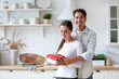 © BestForYou - Woman in a white shirt holding a red gift box with a white ribbon, standing in a kitchen with a man in a light blue shirt behind her.