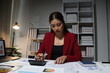 © Wasan - Asian businesswoman working late using calculator and checking reports with charts and graphs in office at night.