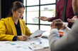© Wasan - Asian businesswoman reviewing document while colleagues discussing project strategy in office, collaboration and data analysis for successful business.
