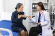 © Wasan - Senior woman sitting on a hospital bed, discussing chest pain symptoms with a doctor, who is taking notes on a clipboard during their consultation in the examination room.