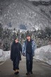 © Creativa Images - ​A couple holding hands walks down a snowy, quiet mountain road in winter, surrounded by snow-covered trees and a steep forest backdrop. They are wearing warm, casual black and gray jackets and sungla