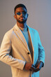 © SHOTPRIME STUDIO - Stylish young man in a beige coat and sunglasses, showcasing a confident expression against a gradient blue background, ideal for fashion and seasonal promotions