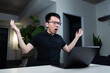 © fotolite - An Asian man wearing glasses raises both hands in a gesture of frustration and confusion while looking at his laptop. He appears exasperated while working late at night in his home office.