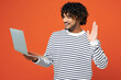 © ViDi Studio - Young happy It Indian man wears striped sweatshirt casual clothes hold use work on laptop pc computer waving hand talk speak isolated on plain red orange background studio portrait. Lifestyle concept.
