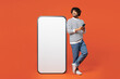 © ViDi Studio - Full body young smiling Indian man wear striped sweatshirt casual clothes stand near big huge blank screen mobile cell phone with mockup area using smartphone isolated on plain red orange background.