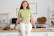 © Pixel-Shot - Smiling young woman in tight jeans sitting on table at home. Weight gain concept