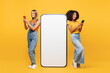 © ViDi Studio - Full body two young friends buddies women wear denim casual clothes together big huge blank screen area mobile cell phone use smartphone isolated on plain yellow orange background. Lifestyle concept.