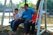 © ROHE Creative Studio - A father relaxing with his two children on a swing at an outdoor recreational area.