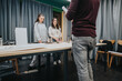 © qunica.com - Two women stand at a table in a collaboration center as a man in the foreground presents. They discuss materials for a project in a focused, collaborative workshop setting.