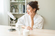© KMPZZZ - Facial beauty skin care, smile of pretty asian young woman in bathrobe looking at mirror, hand applying moisturizer lotion on her face, putting cream treatment before makeup cosmetic routine at home.