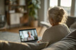 © EricMiguel - Elderly woman video chatting on a laptop at home in a cozy sweater, gray hair in a bun, sunlight illuminating her — comfortable remote connection and care