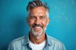 © miss irine - Handsome middle-aged man with grey hair smiles warmly. He wears a blue denim shirt and white t-shirt. His expression is friendly and confident against a solid blue backdrop.