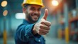 © miss irine - Man in yellow hard hat gives thumbs up gesture. Worker wears blue work clothes inside industrial building, smiling happily. Represents approval or great job.
