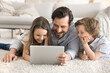 © fizkes - Family portrait with gadget. Loving single dad spend quality time with two little kids daughter son relax on carpet with digital pad watch cartoon play game read ebook search education video content