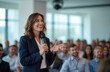 © Viktor - Woman speaks into microphone addressing audience in conference hall. Professional presenter engages listeners with speech at business event. People listen attentively in modern venue.