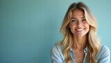 Attractive young blonde woman with blue eyes smiles warmly at camera, indoor studio portrait. Wears casual denim shirt, long wavy hair frames face, soft light features. Confident, serene expression.