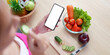 © Natee Meepian - Digital Health. Woman checking smartphone while preparing fresh vegetables, promoting healthy eating.