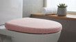 © smth.design - Soft pink fabric toilet seat cover in modern bathroom with neutral tiles, wooden shelf, rolled white towel and small succulent plant