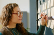 © AnnaStills - Caucasian young adult woman writing on sticky notes attached to glass wall, using pen while organizing tasks, wearing eyeglasses, focusing on project planning in modern workspace