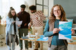 © AnnaStills - Portrait of young adult Caucasian woman smiling at camera holding real estate brochure, while diverse group of young adults discussing documents in modern office background