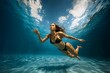 © Loginova - Underwater wide shot of athletic woman in black bikini swimming gracefully through clear blue pool, sunlight patterns dancing on sand-colored bottom.