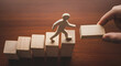 © Pixel Studio - Wooden figure climbs ascending blocks while a hand places the next step symbolizing career growth mentorship and achieving future success goals