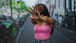 © Krakenimages.com - Woman shielding face with splayed hands showing long nails while wearing pink tanktop and jeans on a city street lined with parked bicycles and buildings; privacy unease.