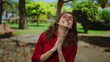© Krakenimages.com - Woman in red shirt smiling joyfully with hands together, surrounded by lush park greenery on a sunny day, conveying happiness and serenity in an outdoor setting.