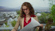© Krakenimages.com - Woman in red shirt and glasses examines blueprints outdoors in a city setting with modern architecture and green landscape in the background.