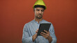© Krakenimages.com - Young hispanic man in hardhat uses tablet against isolated red background, suggesting modern construction technology with focused expression.