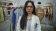 © Krakenimages.com - Young woman scientist in white uniform standing in a clothing store with racks and outfits in background, wearing safety goggles and focusing indoors on her work environment.