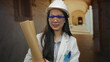 © Krakenimages.com - Young chinese woman architect outdoors in old town holding blueprints, wearing a hardhat and glasses, with a smile, showcasing her profession in a charming historical setting.
