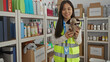 © Krakenimages.com - Woman in reflective vest holding stuffed animal in charity donation center filled with boxes and hygiene products, showcasing volunteer work in indoor setting.