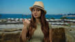 © Krakenimages.com - Woman in straw hat pointing thumb at scenic beach port with clear sky, showcasing young hispanic travel vibe on a sunny day outdoors, with boats in the background.