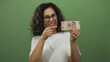 © Krakenimages.com - Middle-aged hispanic woman with glasses smiles holding mexican pesos against a green isolated wall, emphasizing a financial theme.